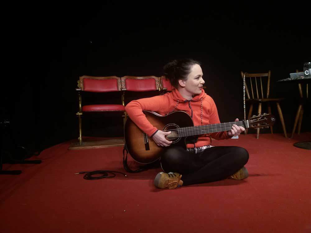 Katrin Freiburghaus auf dem Boden sitzend, dabei eine Gitarre spielen. Foto von Fee Brembeck.