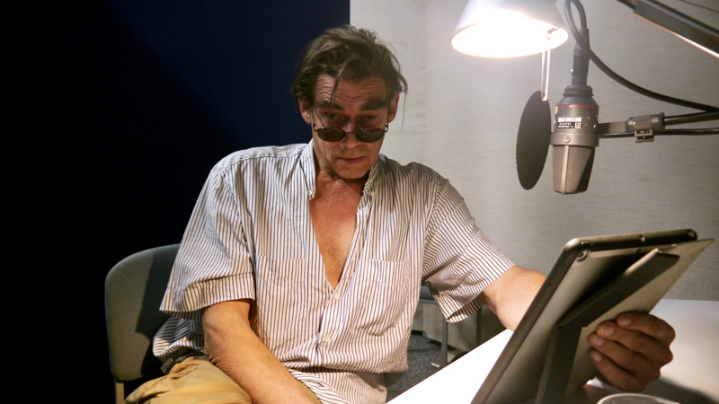 Ein Mann mit Sonnenbrille, geöffnetem Hemd sitzt vor einem Mikrofon mit Lampe und blickt in die Kamera: Martin Feifel, spricht ein für das BR-Hörspiel "Blut".