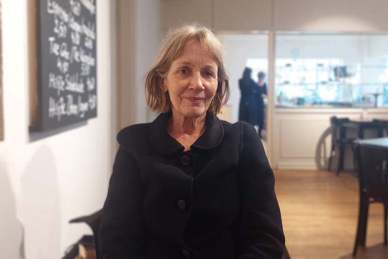 Eine ältere Frau im schwarzen Mantel sitzend in einem Café und in die Kamera lächelnd: Dagmar Leupold.