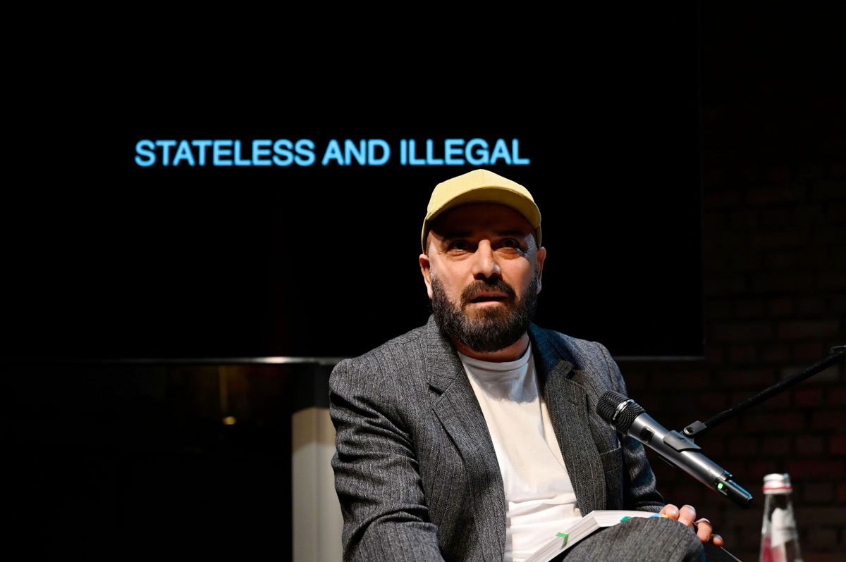 Delschad Numan Khorschid liest im Rahmen des Welt/Bühne-Salons im Marstalll Residenztheater, hinter ihm Text: Stateless and illegal