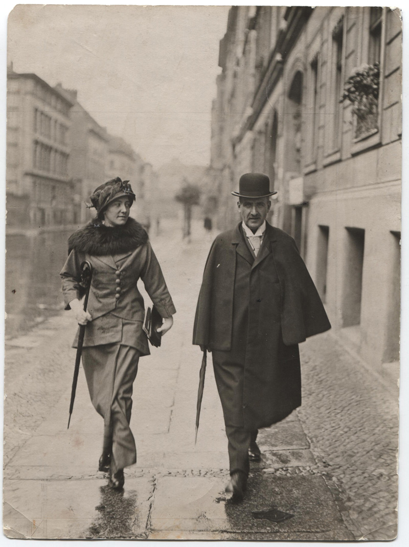 Historisches Foto: Mann und Frau mit Regenschirm in der Hand blicken teils in die Kamera und gehen eine Straße entlang: Tilly und Frank Wedekind.