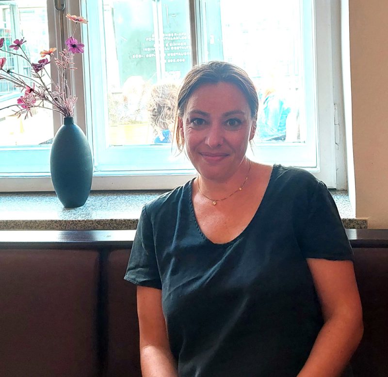 Frau vor einem Fenster sitzend mit Blumenvase im Hintergrund, lächelnd in Kamera blickend: Joana Osman.