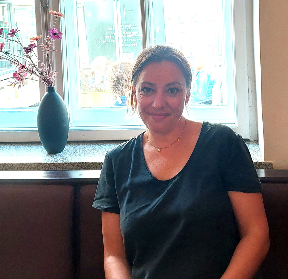 Frau vor einem Fenster sitzend mit Blumenvase im Hintergrund, lächelnd in Kamera blickend: Joana Osman.