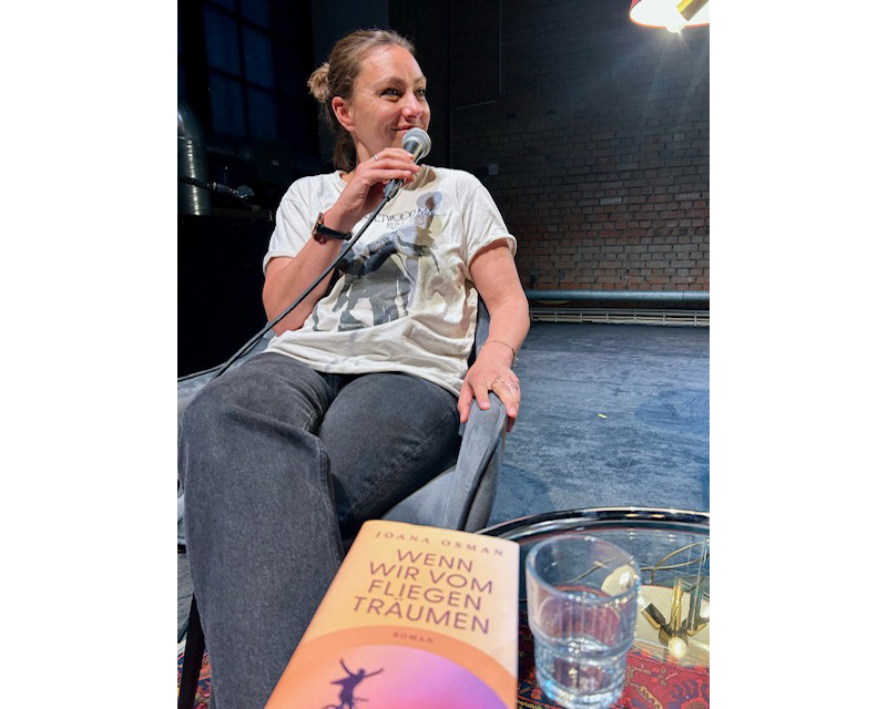 Frau auf Bühne sitzend mit Mikrofon nach rechts blickend, im Vordergrund Buch "Wenn wir vom Fliegen träumen": Joana Osman.
