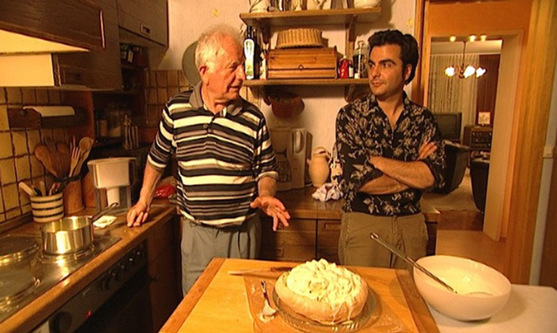 Karnik als junger Mann und sein Vater im Gespräch in der Küche hinter einem traditionellen Brot-Gericht aus zerrupftem Brot, das mit Joghurt und zerlassener Butter geschichtet ist.