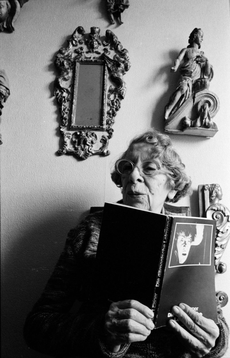 Porträt ältere Dame mit Brille, liest im aufgeschlagenen Buch, an Wand Spiegel und Engeldarstellungen.