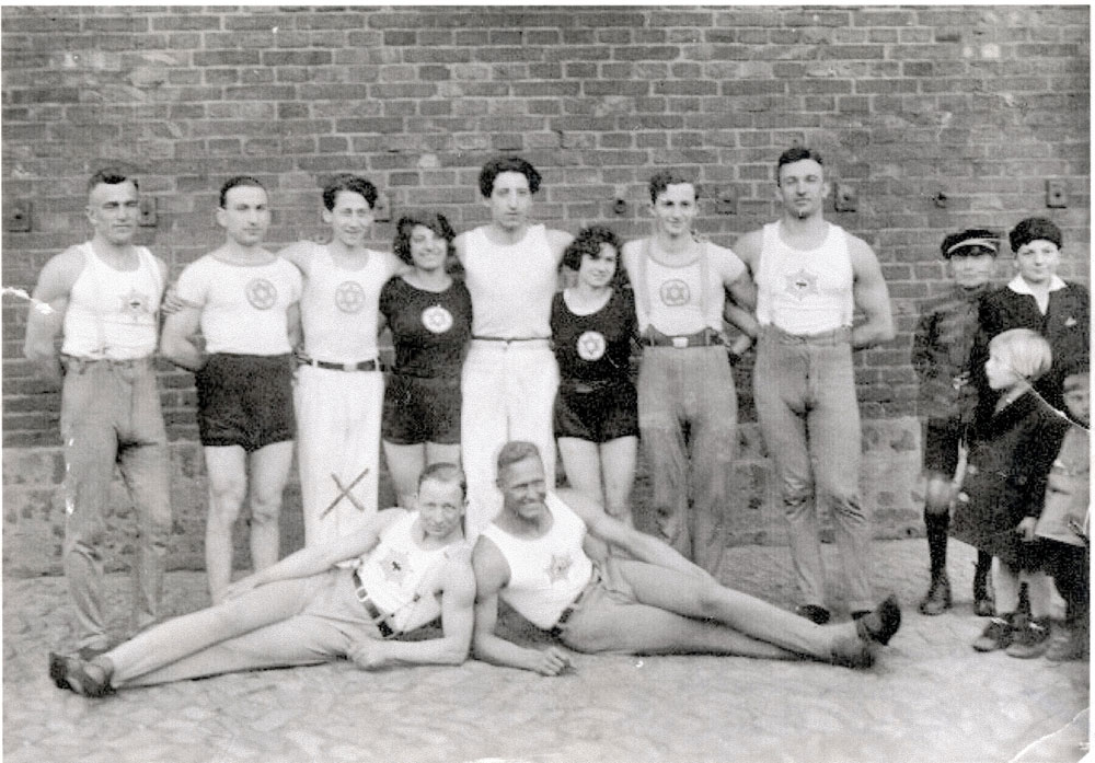 Sportler und Sportlerinnen stehen vor Hausfassade, zwei liegen vor ihnen rechts blicken Kinder zu ihnen - schwarz-weiß-Fotografie