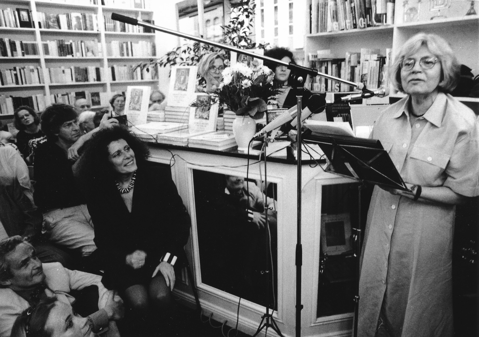 Schwarz-weiß-Foto einer Veranstaltung in einer Buchhandlung, eine ältere Frau mit Brille steht vor einem Mikrofon, vor ihr sitzen Menschen und lauschen, darunter neben Ruth Gay sitzt Rachel Salamander.