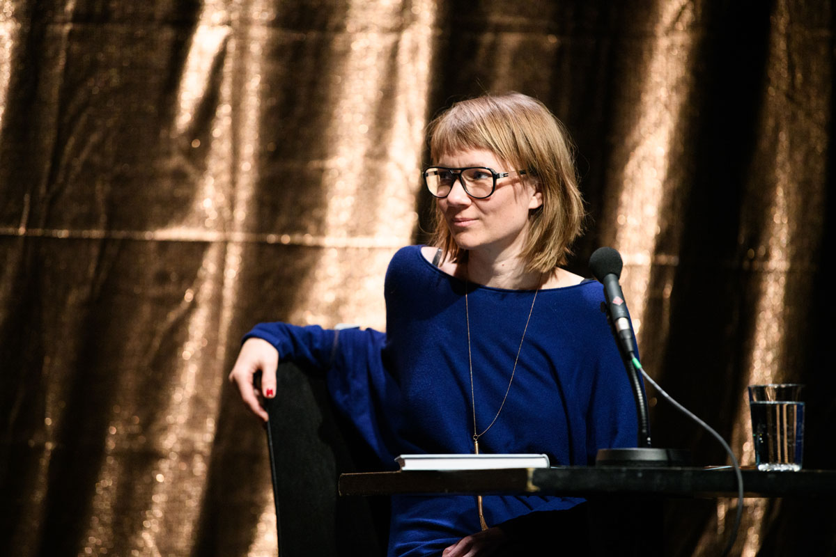 Raphael Bardutzky vor goldenne Vorhang im blauen Pulli auf Podium sitzend. Sie moderiert LIX