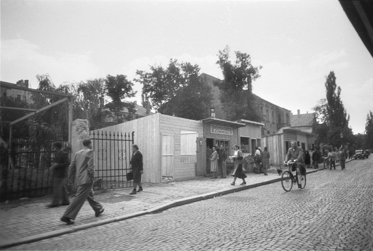 Gepflasterte Straße mit Geschäften in Bretterbuden auf Gartengrundstücken. Schwarzmarkt in der Möhlstraße in Bogenhausen. Personen beim Einkauf zu Fuß und auf dem Fahrrad.