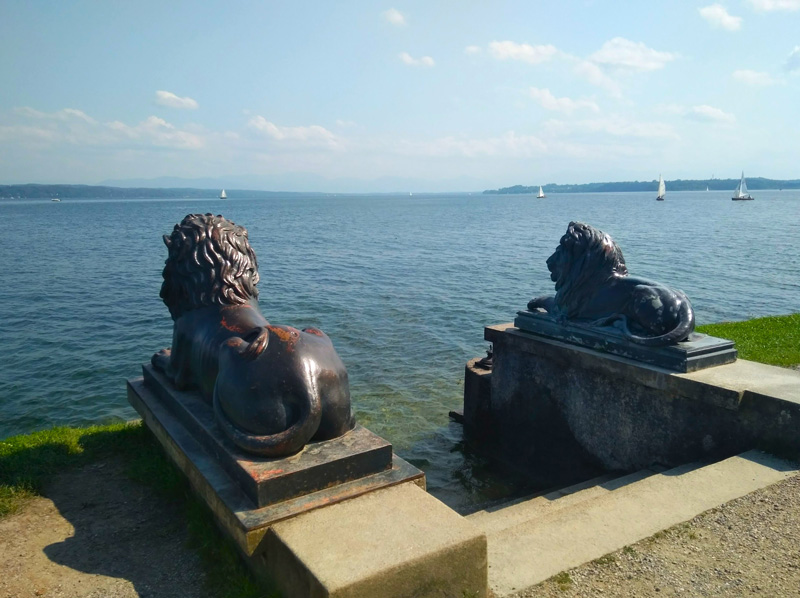 Zwei bronzene Löwe an Treppe mit Blick auf einen See mit Segelbooten.