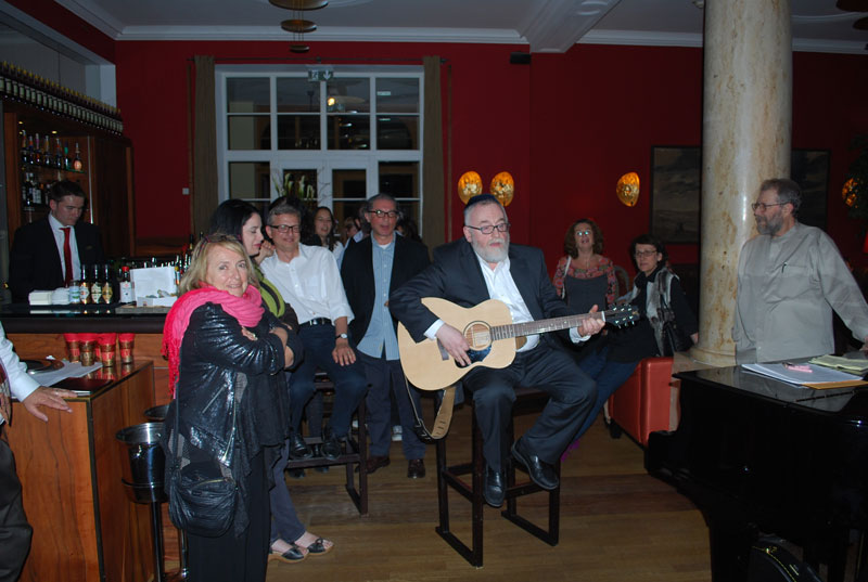 Oberrabiner Paul Chaim Eisenberg sitzt auf Hocker und spielt Guitarre vor Publikum in einer Bar mit Piano. Tarbut 2012.