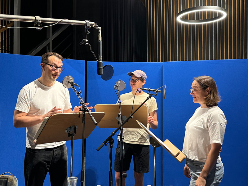 Drei Personen stehen vor blauem Hintergrund und sprechen in Mikrofone: BR-Hörspiel "Nie wieder gut" mit Steven Scharf, Vincent Sauer und Judith Toth.