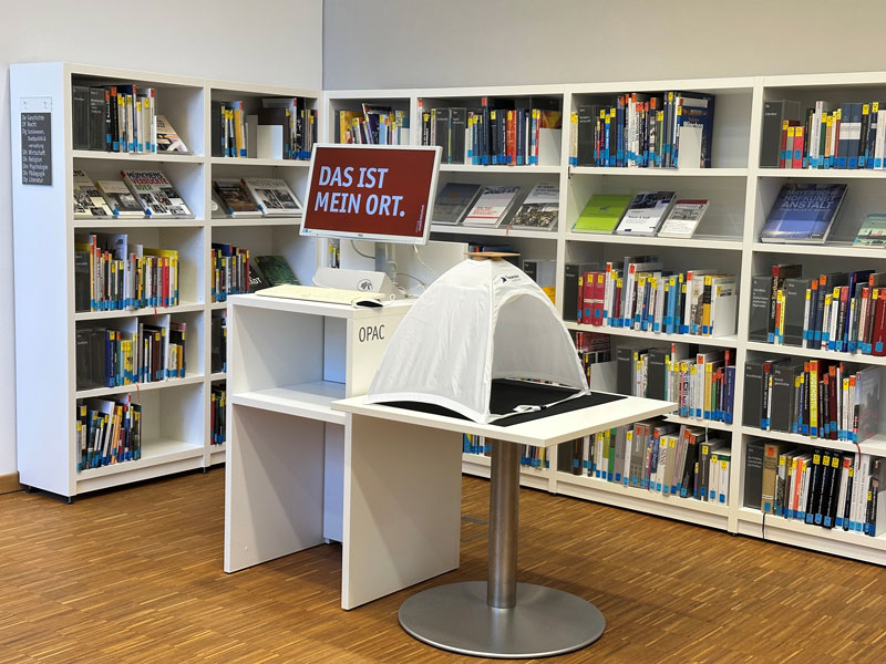 Blick auf gefüllte Bücherregale mit Computer davor für den Opac und dem Screen "Das ist mein Ort".