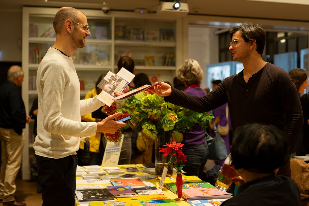 zwei Männer am Tisch stehend, die sich Broschüren übergeben. FemaleHeritage, Open House der freien Szene.