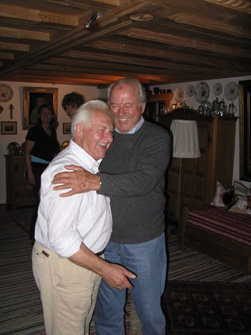 zwei Männer lachend, stehend, der jüngere umfasst den kleineren älteren in einer Stube: Franz Westermeier und sein Sohn.