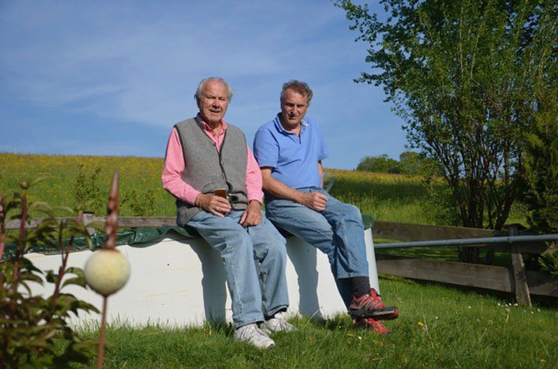 zwei Männer unterschiedlichen Alters blicken zur Kamera auf einem Mauervorsprung in Natur sitzend: Franz und Sebastian Westermeier.