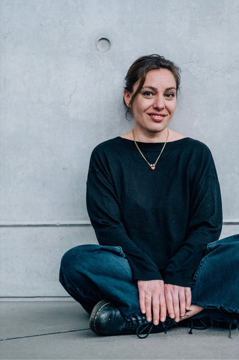 Frau im Schneidersitz vor Wand, lächelnd, dunkel gekleidet mit zusammengebundenen Haaren: Joana Osman.