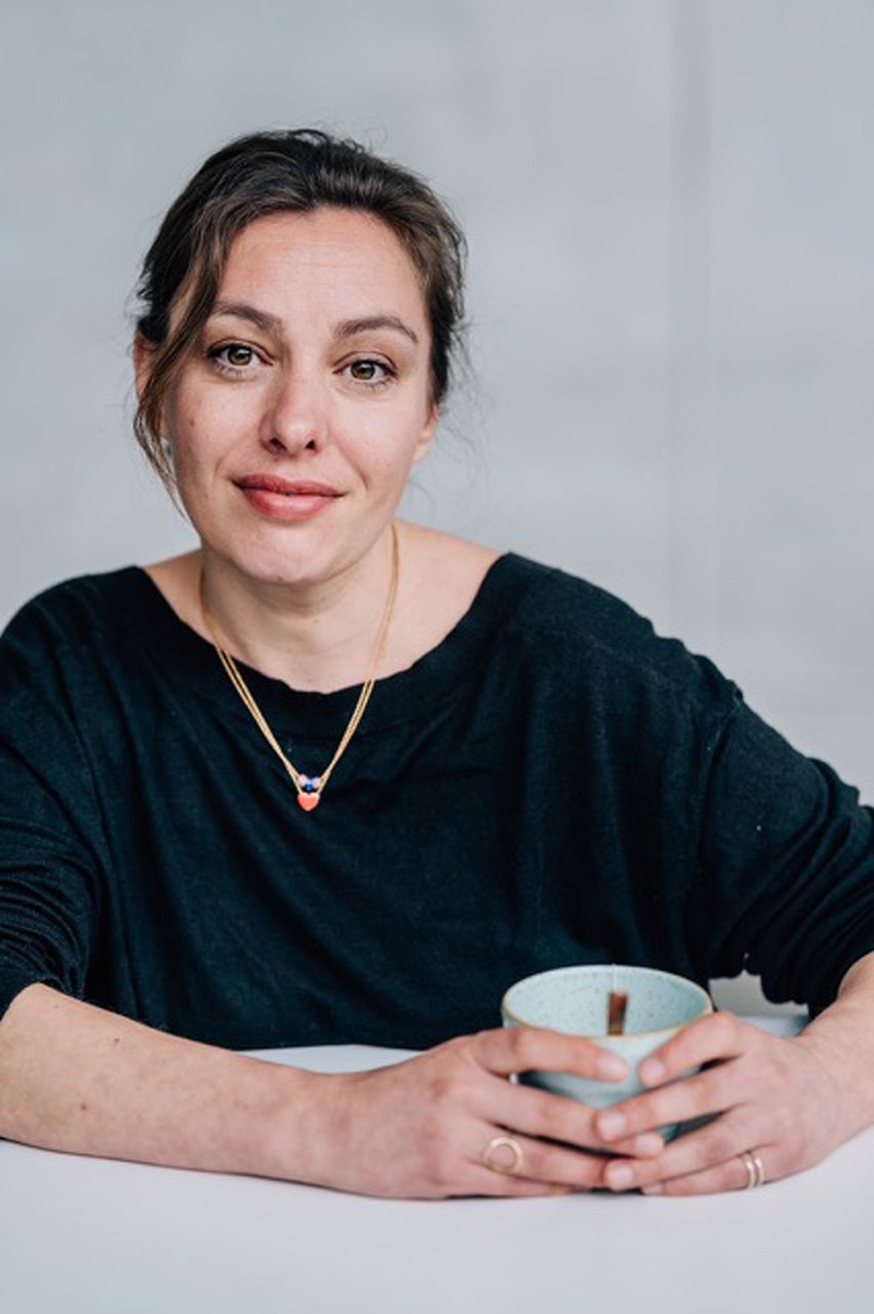 Frau am Tisch sitzend mit einer Teetasse in der Hand, zusammengebundene Haare, in Kamera blickend: Joana Osman.