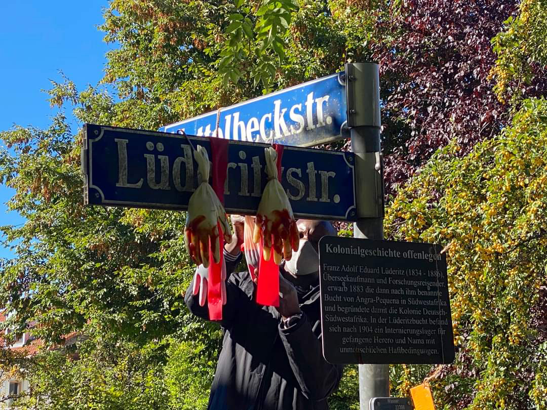 Mann hängt „blutverschmierte“ Handschuhe am Straßenschild Lüderitzstraße, Ecker Nettelbeckstraße auf. Auf den Spuren von kolonialen Straßennamen mit Hamado Dipama.