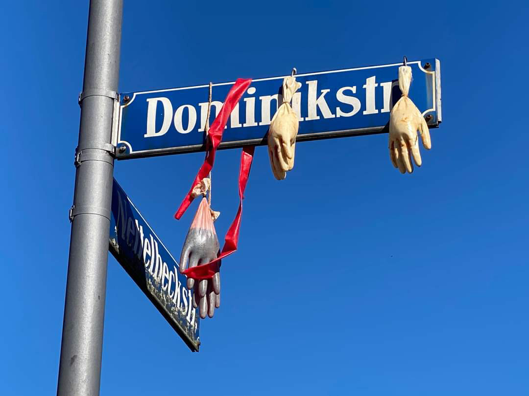 Mit "blutgefüllte" Handschuhe hängend am Straßenschild Dominikstr. in München. Aktion gegen koloniale Straßennahmen