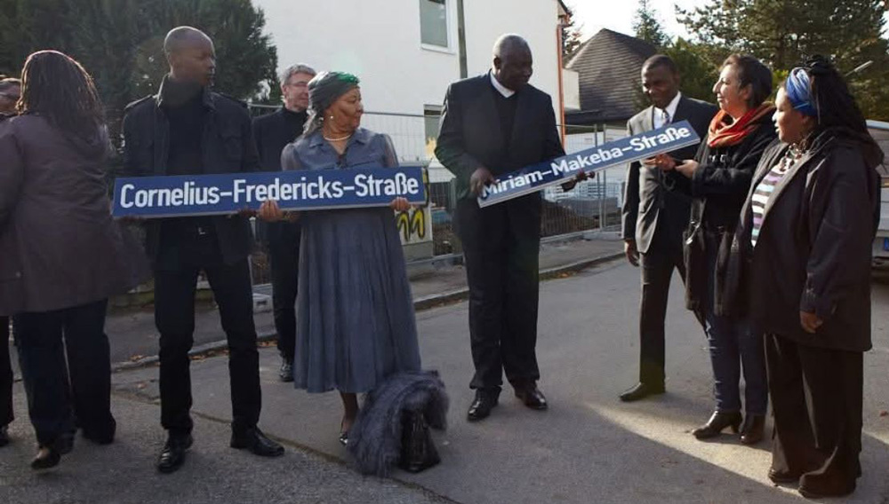 Vier Personen halten zwei symbolische Straßenschilder mit Aufschrift Cornelius-Fredericks-Straße und Miriam-Makeba-Straße in Bogenhausen. Hamado Dipama und koloniale Straßennamen in München-Bogenhausen.