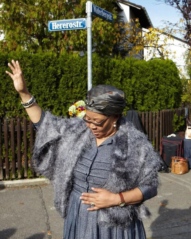 Frau mit erhobener Hand vor dem Straßenschild Hererostr. in München Bogenhausen. Auf den Spuren kolonialer Straßennamen.