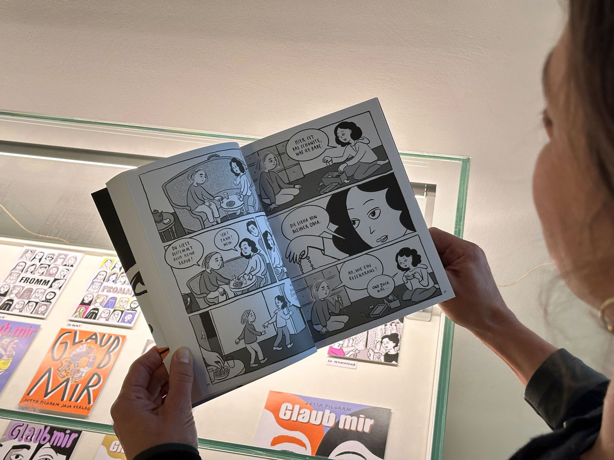 Frau hat über eine Vitrine ein Comic aufgeschlagen. In der Vitrine sieht man mehrer bunte Skizzen des Comics, eine davon mit dem Titelbild des Comic «Glaub mir» von Jutta Pilgram.