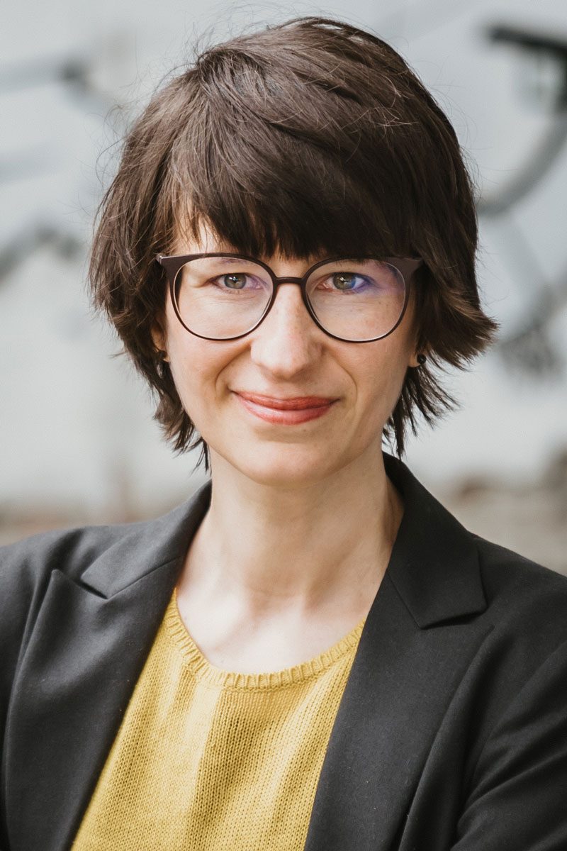 Frauen-Porträt, kurze dunkle Haare mit dunkler Brille, schwarzem Jacket und gelben Pullover blickt lächelnd in die Kamera: Lisa Frühbeis.