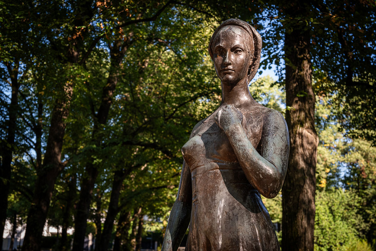 Ansicht auf eine bronzene Frauenskulptur, die im Grünen steht und sich ans Dekolleté greift. Literarischer Spaziergang zu #MeinBogenhausen