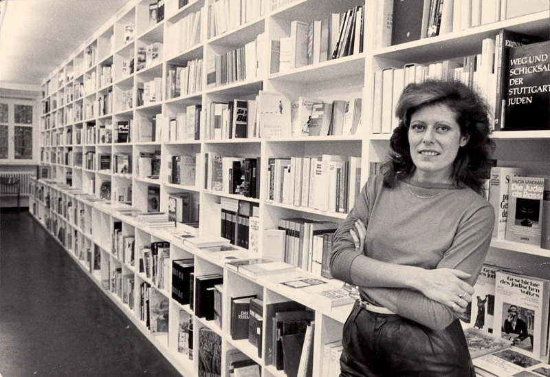Frau mit verschränkten Armen vor einem langen Bücherregal: Rachel Salamander in der Literaturhandlung. Ausstellung in der Monacensia ab 20. Mai 2026.