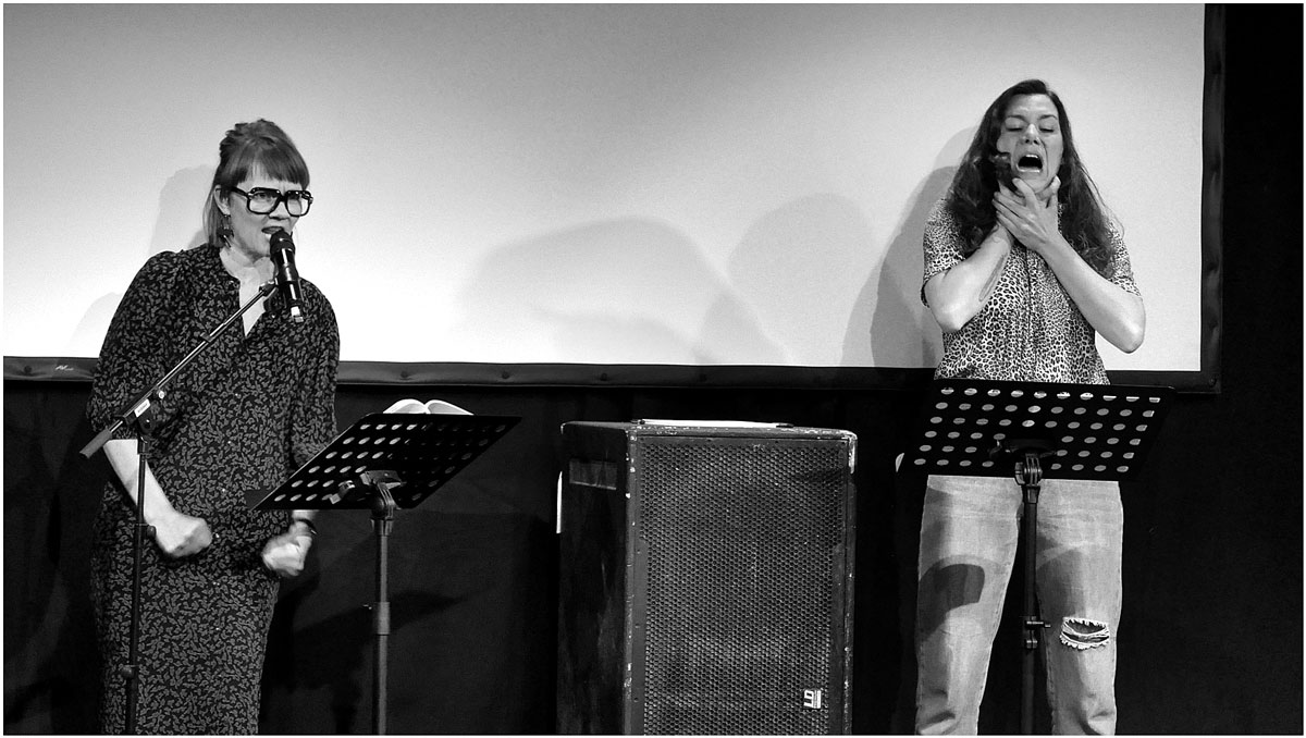 Raphala Bardutzky am Mikrofon auf Bühne, daneben Athena Lange, die schreit und sich an den Hals greift. Theaterstück "Altbau in zentraler Lage", aufgeführt zur Münchner Lesereihe GLITSH.
