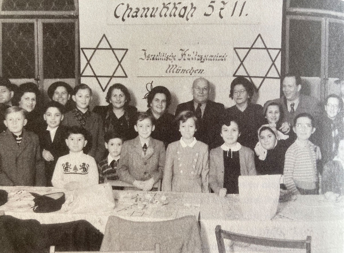 Chanukka-Feier der Israelitischen Kultusgemeinde München von 1950. Kinder und Erwachsener an Tisch in Reihen gestellt, darunter auch Roman Haller mit Löwenpullover.