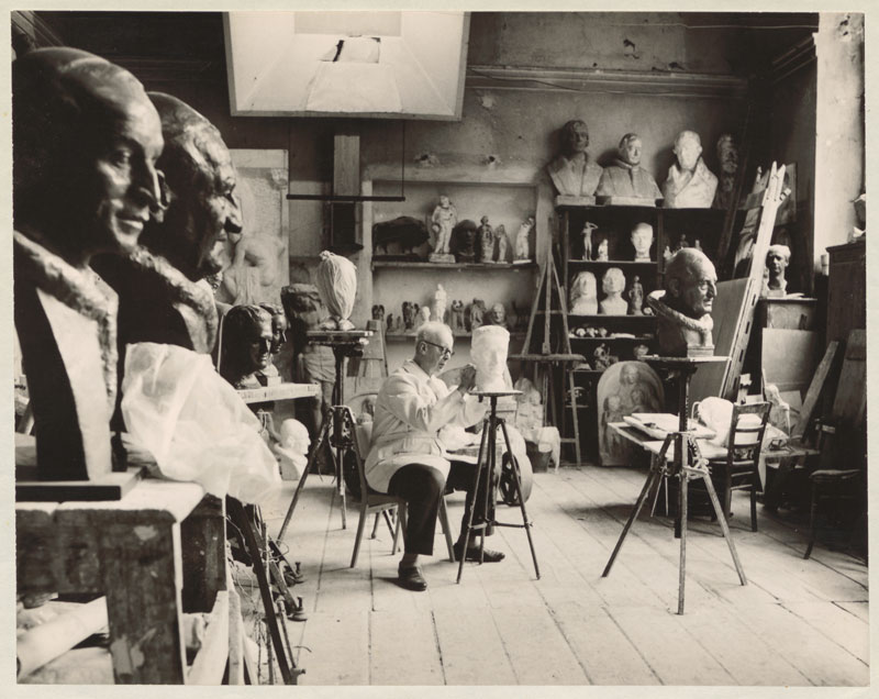 Theodor Georgii sitzt auf Stuhl im Atelier und bearbeitet einen Kopf, im Hintergrund lauter Büsten.