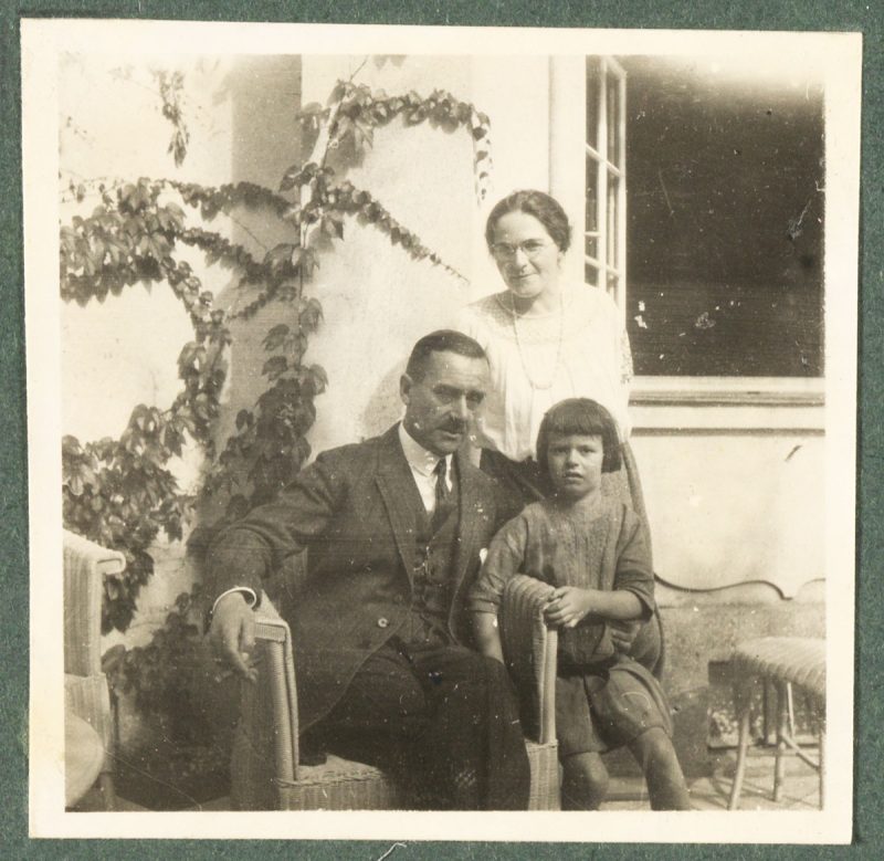 Thomas Mann in einem Korbstuhl sitzend, den linken Arm um Elisabeth Mann gelegt, die neben ihm steht, hinter ihnen stehend Ida Herz. Aus einem Familienalbum.