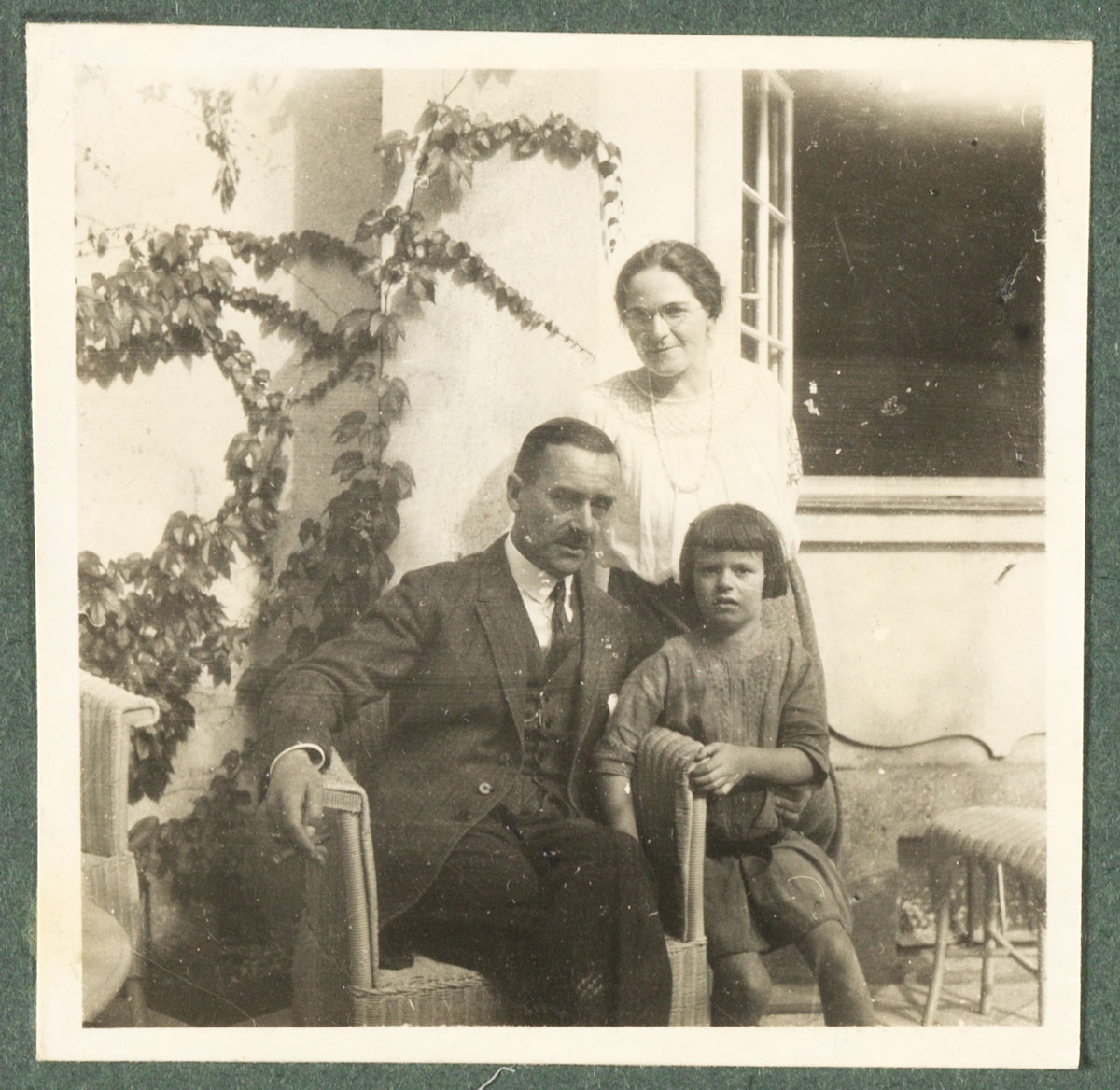 Thomas Mann in einem Korbstuhl sitzend, den linken Arm um Elisabeth Mann gelegt, die neben ihm steht, hinter ihnen stehend Ida Herz. Aus einem Familienalbum.