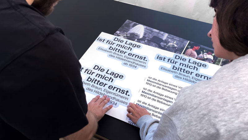 zwei Gestalter von Büro Alba beugen sich über Texttafeln der Ausstellung "Maria Theresia 23. Biografie einer Münchner Villa" - Thumbnail zum Gestaltungsvideo auf Youtube